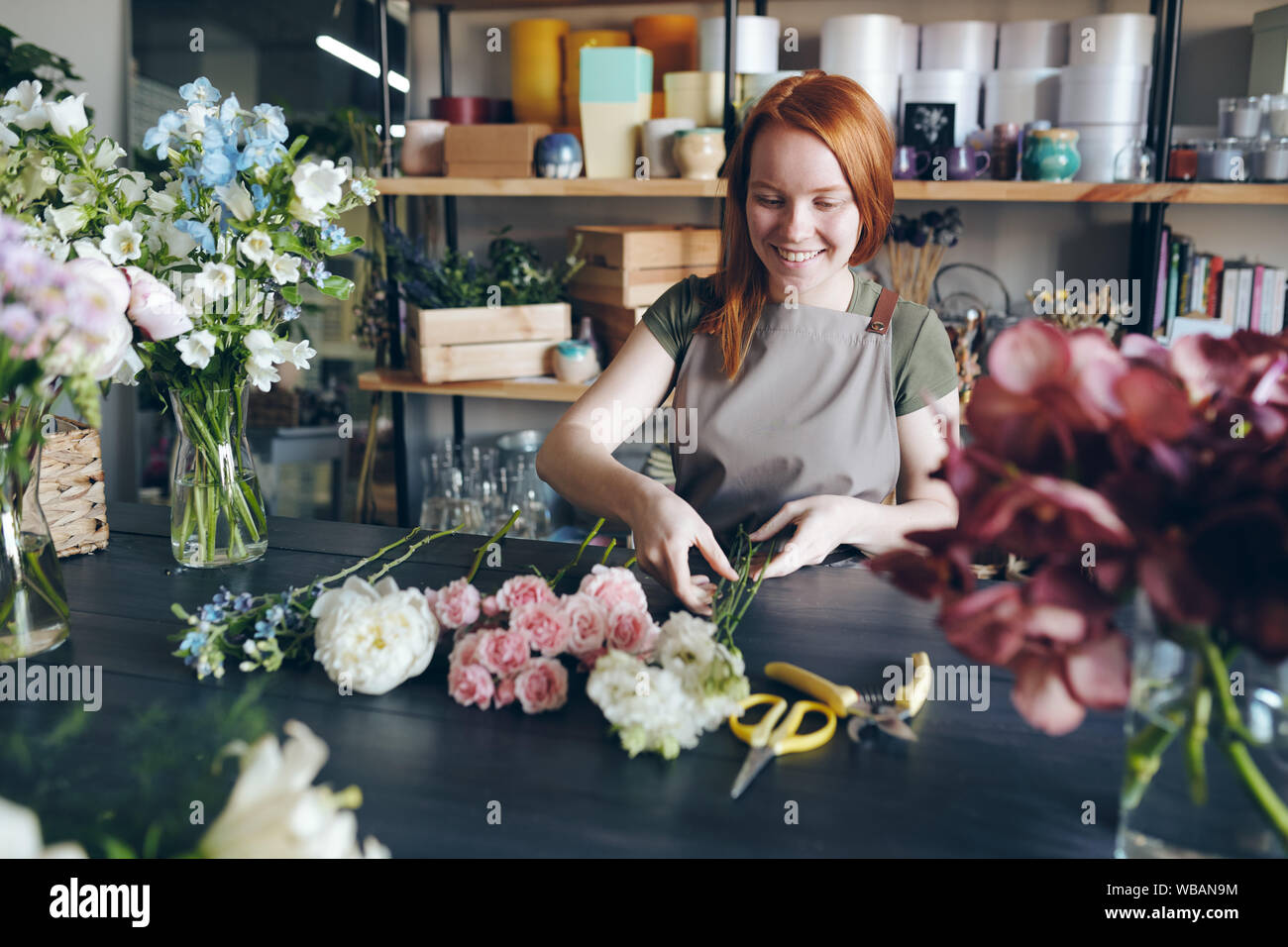 Cheerful beautiful redhead florist in apron standing at table Stock Photo
