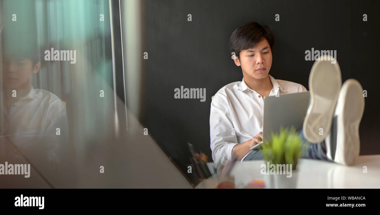 Young male freelancer put his feet on the table while working on a laptop computer in trendy work space Stock Photo
