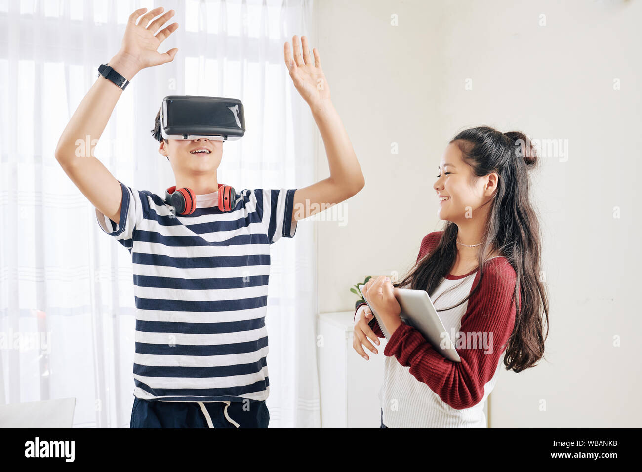 Smiling mixed race teenage girl looking at her brother playing video game in virual reality headset Stock Photo