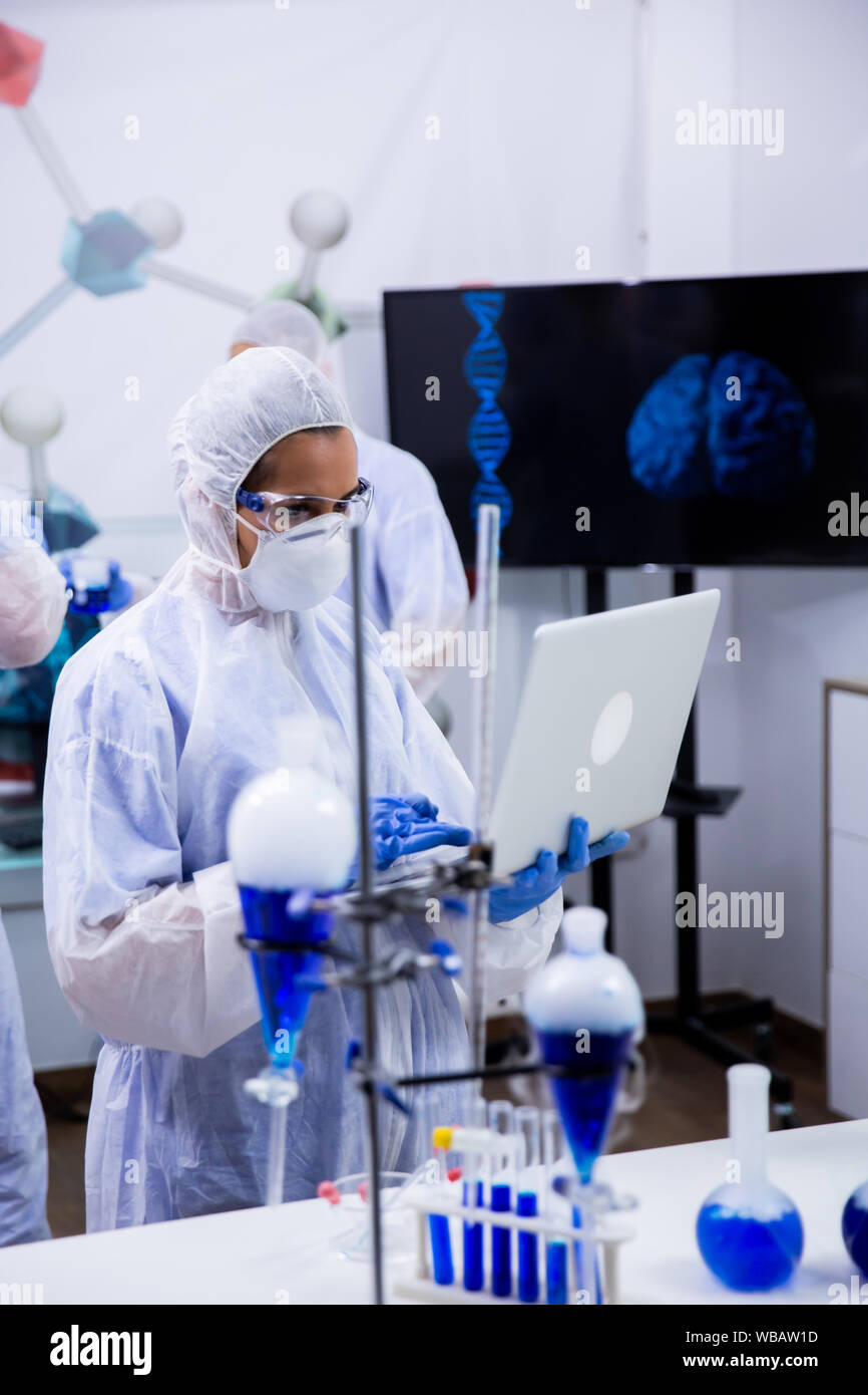 Scientist typing on her computer data from exeperiments with smoking blue solution in test tubes. Stock Photo