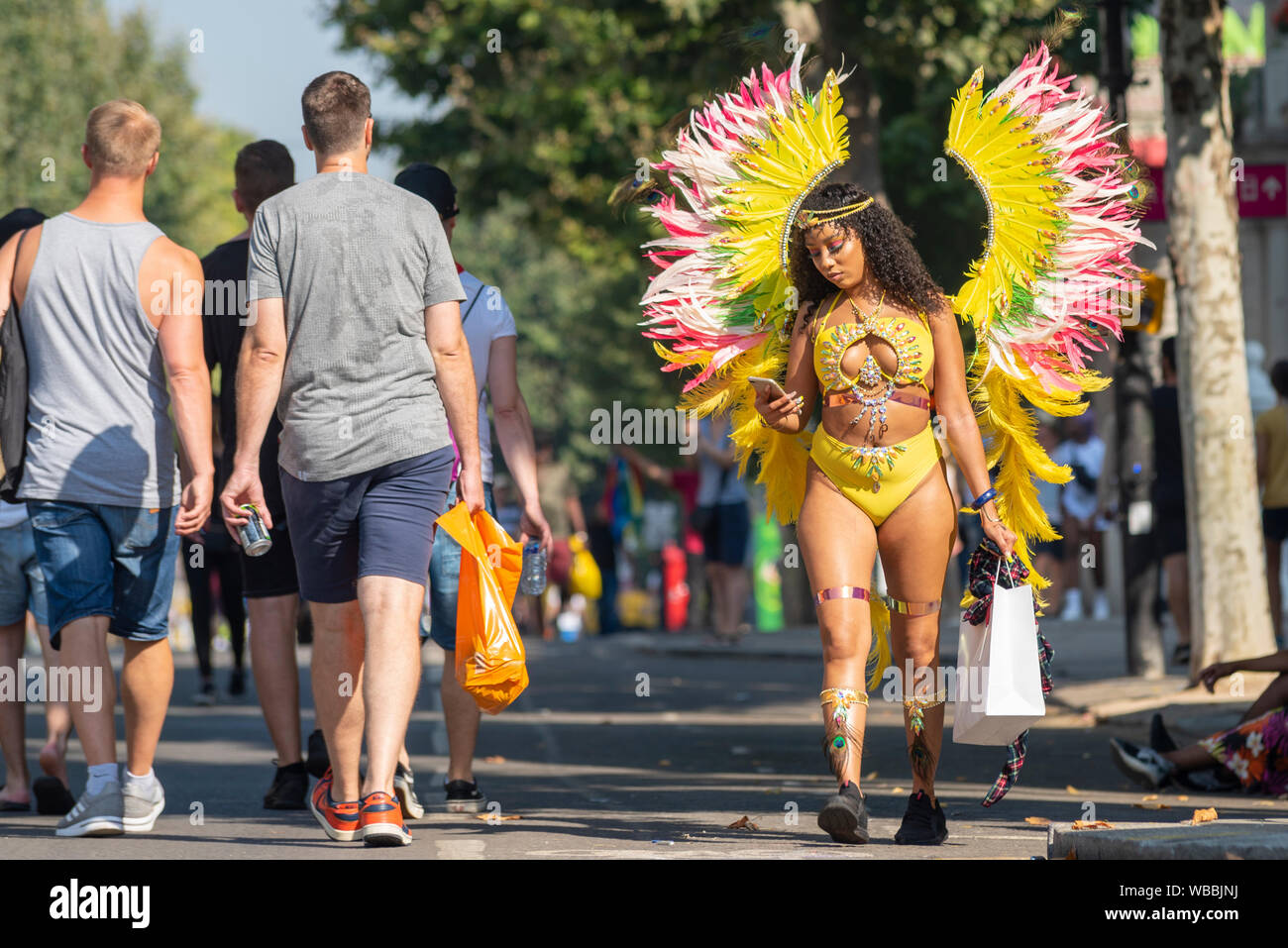 Notting Hill Carnival Parade female participant arriving, wandering past people in brightly coloured extravagant angel wings costume oblivious to her surroundings looking at her phone. Out of place Stock Photo