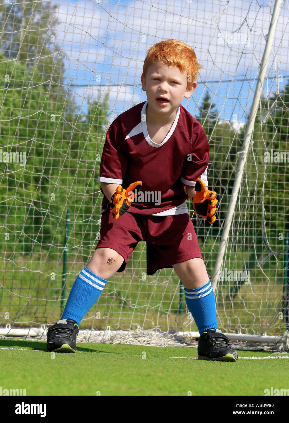 Goalkeeper kid is waiting to catch a ball from a penalty kick on a soccer stadium. Cheerful little boy with ginger hair. Footballer in sports clothes Stock Photo
