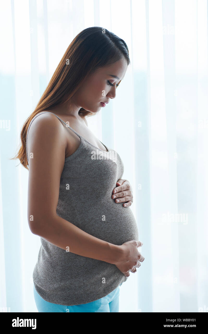 Calm young Vietnamese pregnant woman standing against window and