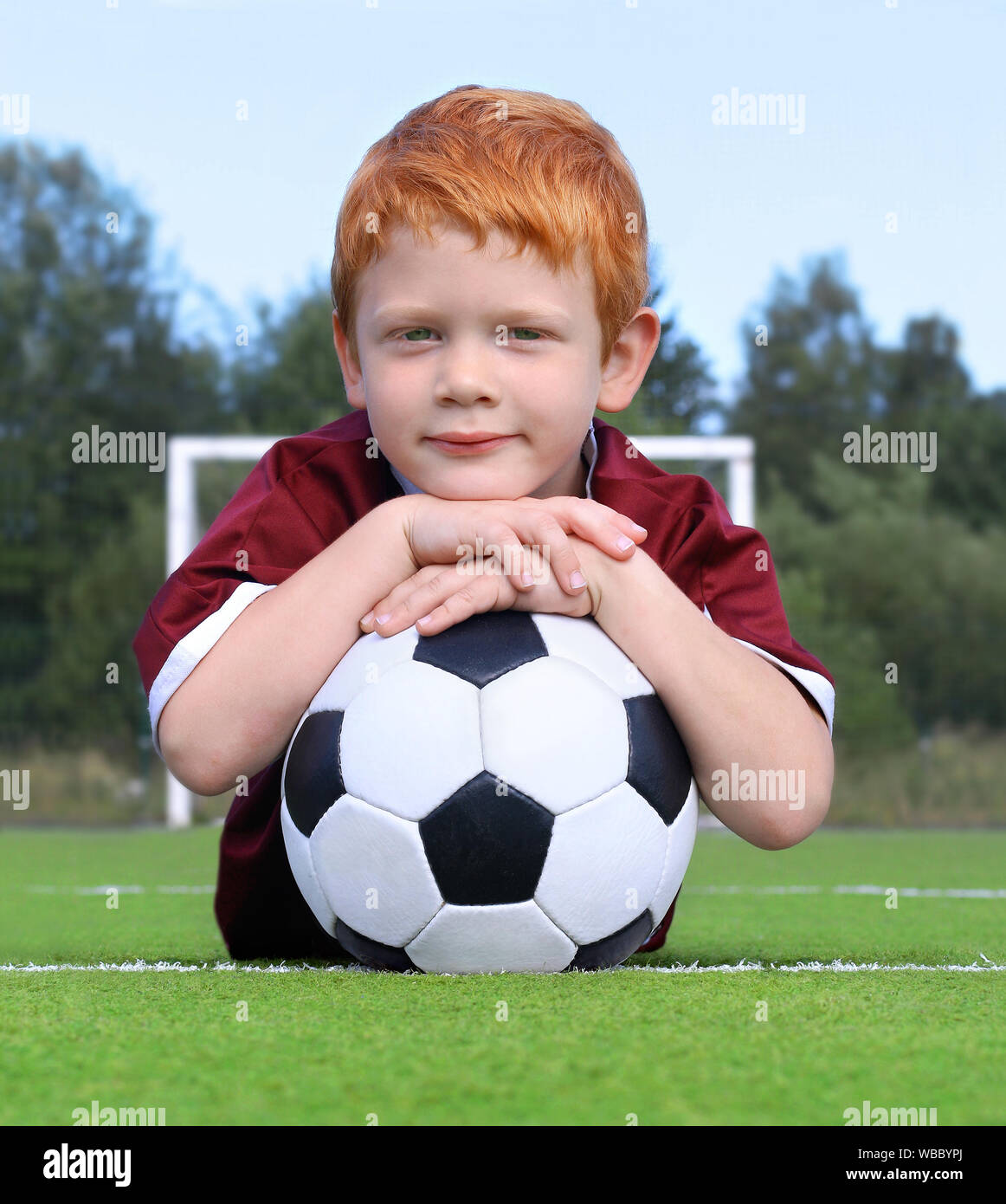 Cheerful little boy with ginger hair posing with a soccer ball. Portrait of a kid in sportswear. Little boy lying on the green lawn. Soccer champion Stock Photo