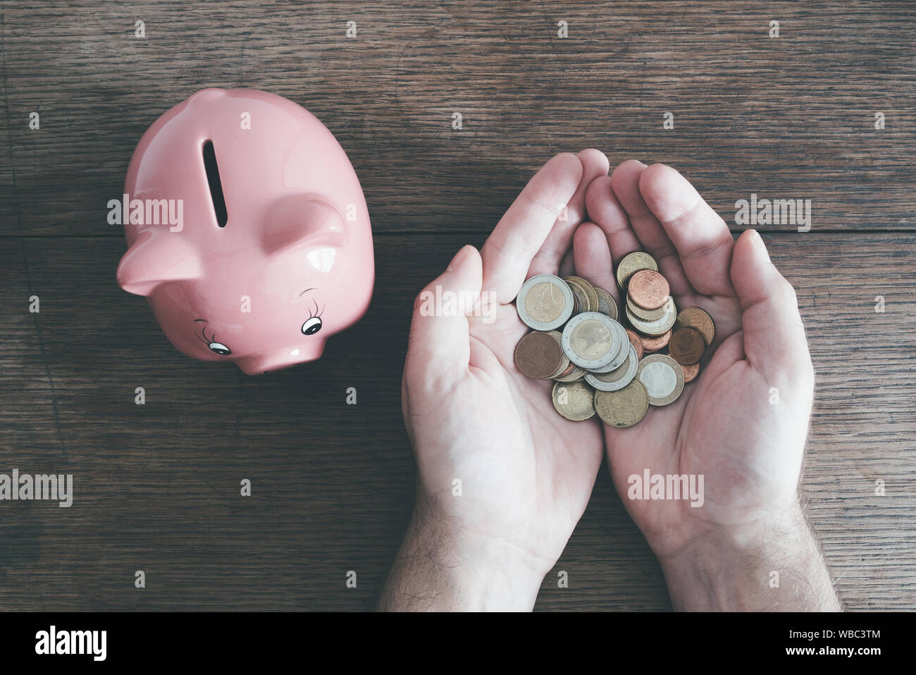 top view of hands of man holding coins next to piggy bank on rustic wooden table, saving money concept Stock Photo