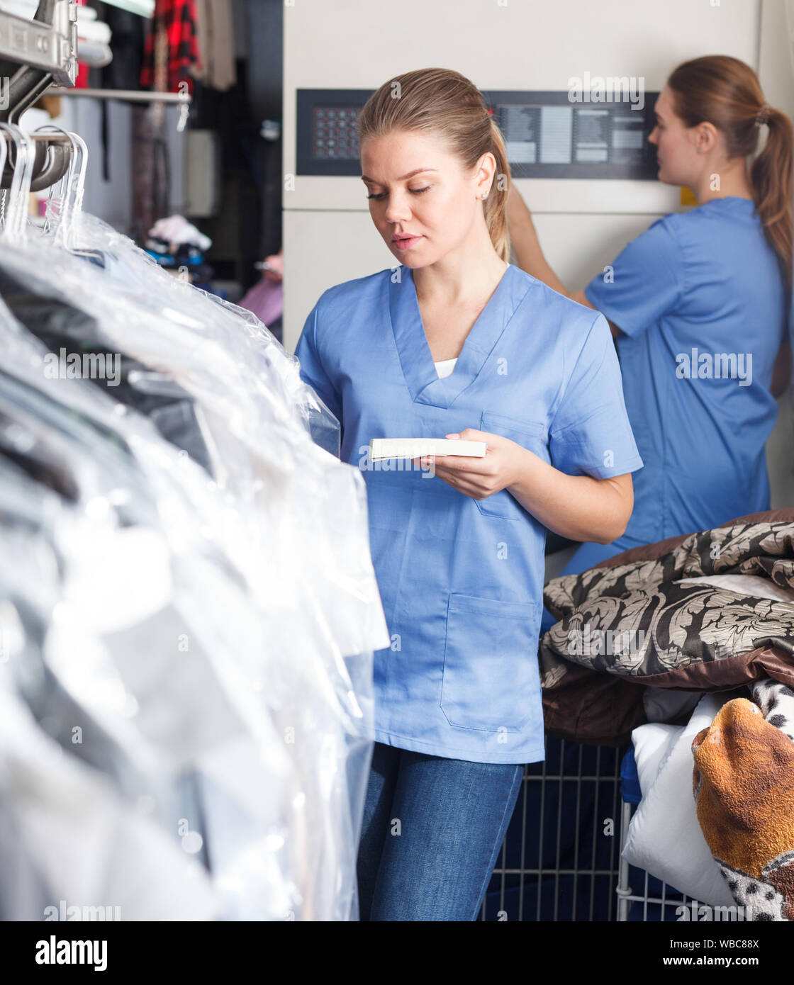 Young female worker of laundry inspecting clothing after dry cleaning on racks Stock Photo