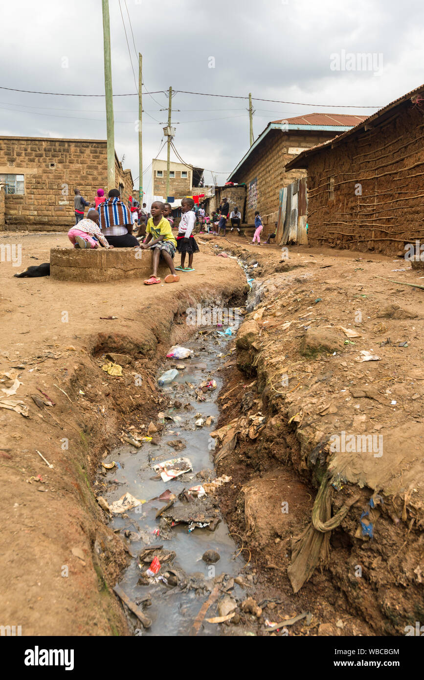 An open sewer flowing between buildings with children and adults beside it, Korogocho slum, Nairobi, Kenya Stock Photo