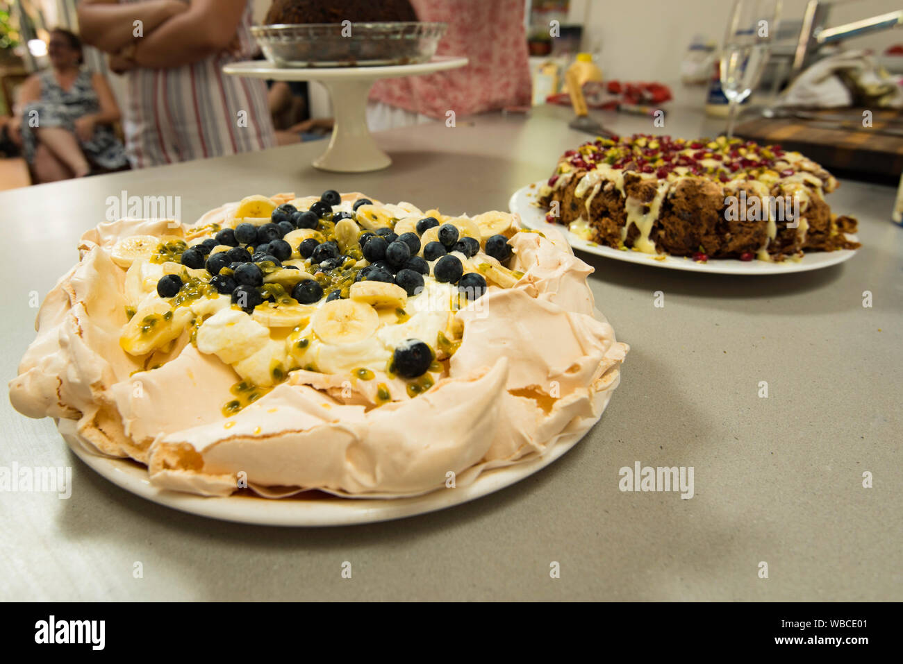 An Australian home made Pavlova dessert covered in cream, blue berries, banana and passionfruit with an iced Christmas cake and pudding to the rear Stock Photo