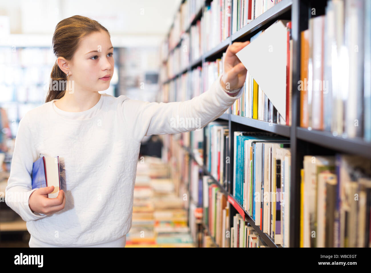 preteen schoolgirl standing in library with pile of books in hands ...