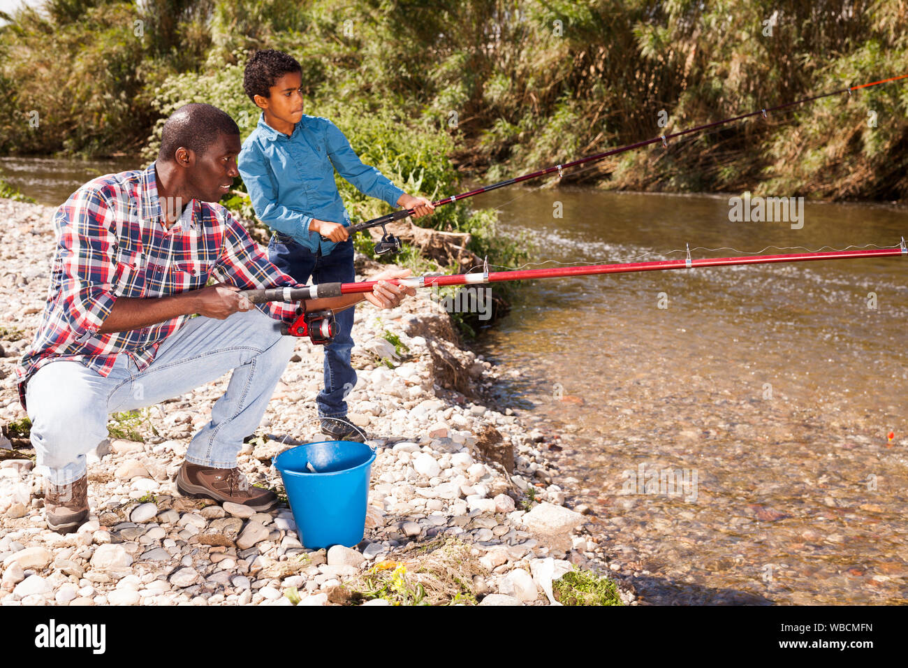 Portrait of cheerful afro man and his little son fishing with rods on river Stock Photo