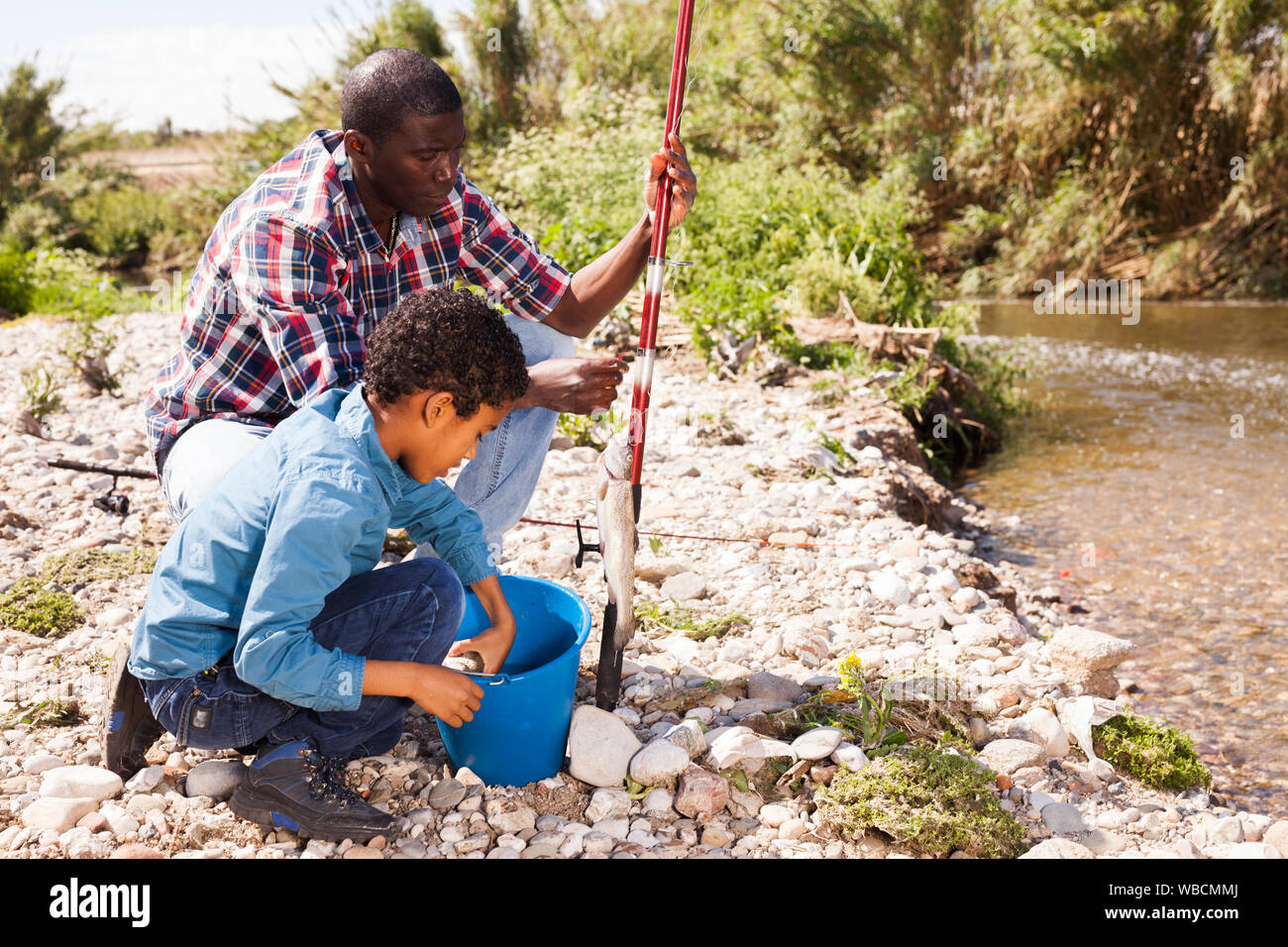 African man and little boy standing near river and holding fish on fishing rod Stock Photo