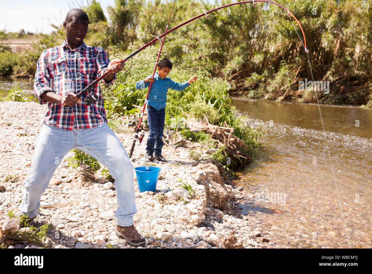 Portrait of Afro man pulling fish with fishing rod and his son fishing on river Stock Photo
