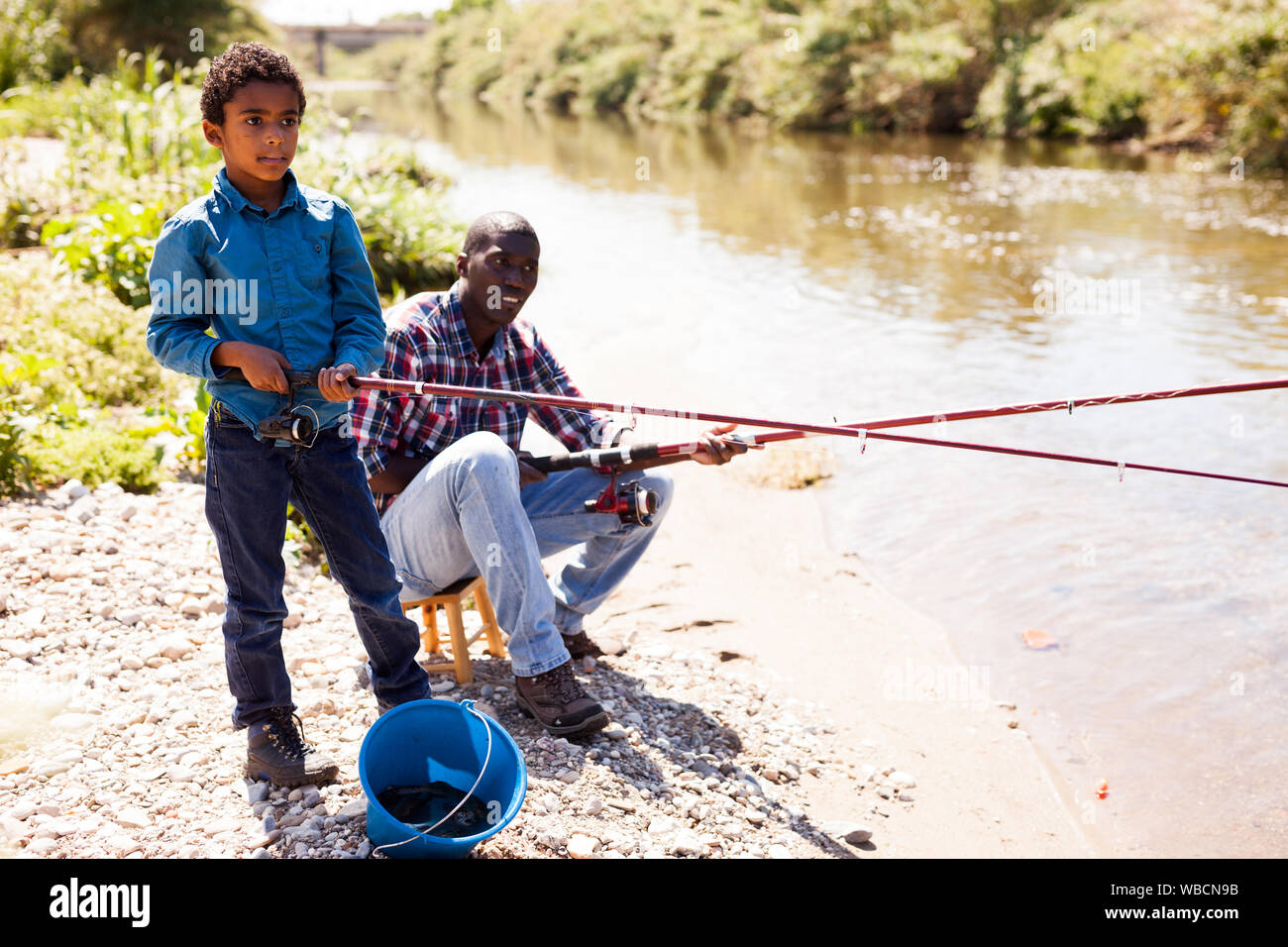 Positive African man and little boy standing near river and fishing with rods Stock Photo