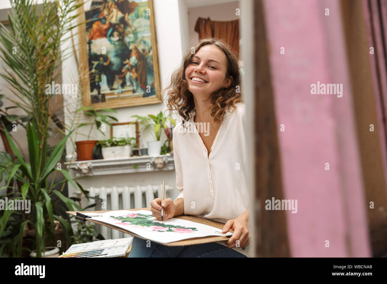 Portrait of affable artistic woman sitting on chair while drawing picture on paper with pain brush and acrylic colors in workshop or master class Stock Photo