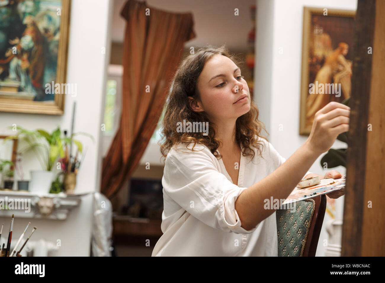 Image of beautiful artistic woman sitting on chair while drawing picture on canvas with pain brush and acrylic colors in workshop or master class Stock Photo