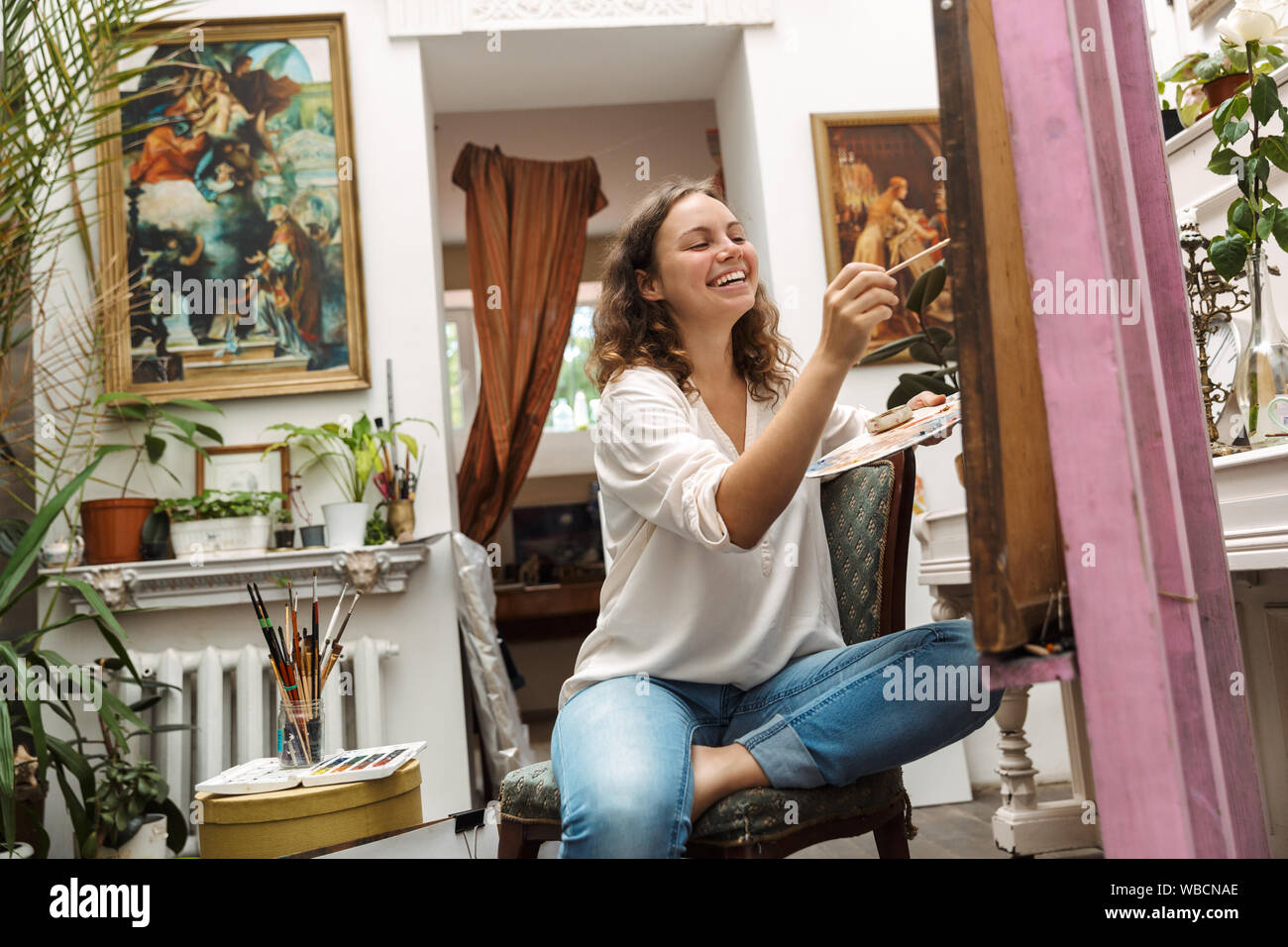 Image of smiling artistic woman sitting on chair while drawing picture on canvas with pain brush and acrylic colors in workshop or master class Stock Photo