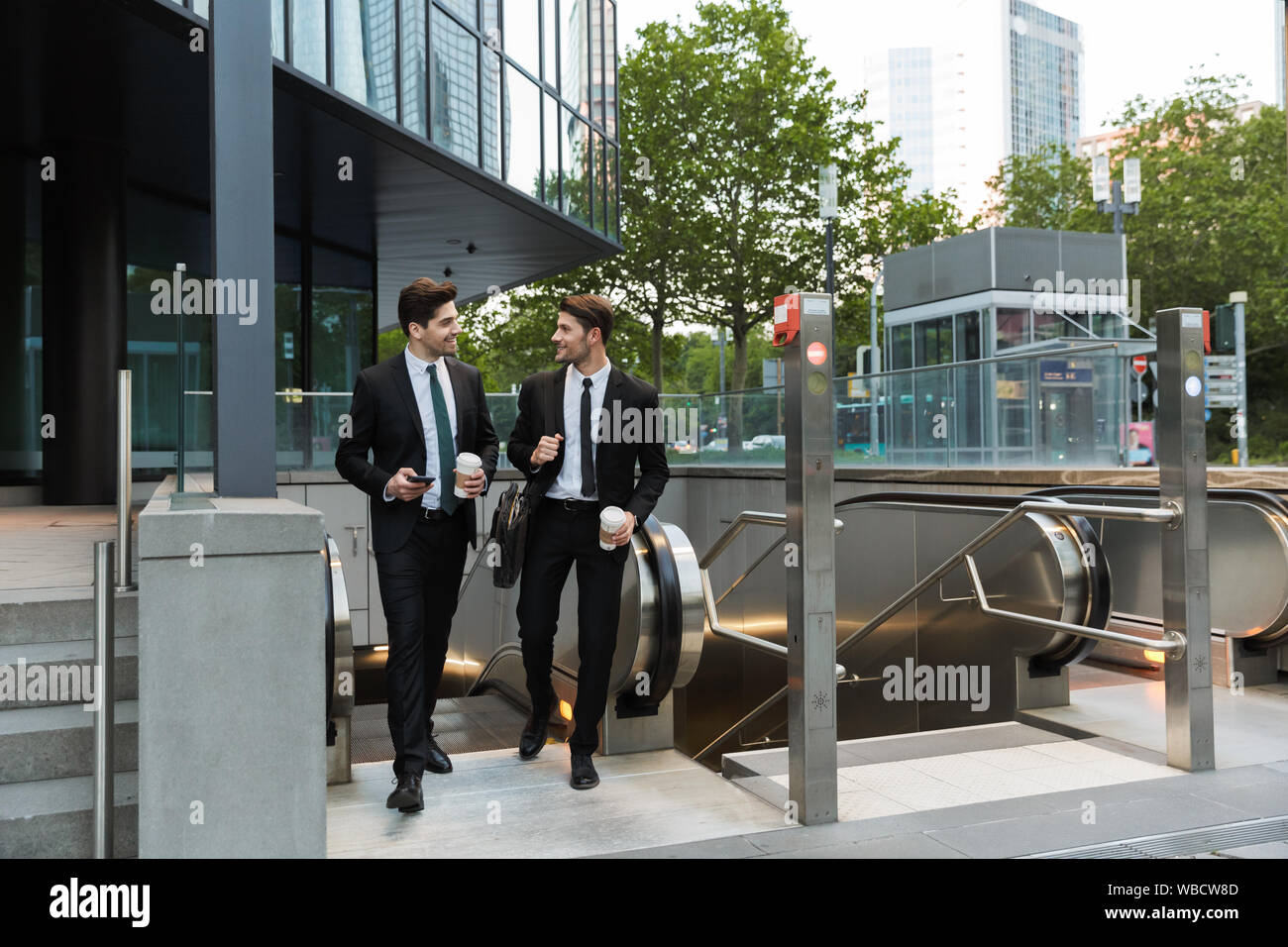 Two attractive confident young businessmen wearing suits walking outdoors at the city streets, having cup of coffee Stock Photo