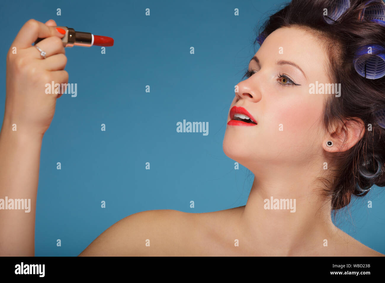 Young woman preparing to party, girl styling hair with curlers, applying makeup red lipstick retro style blue background Stock Photo
