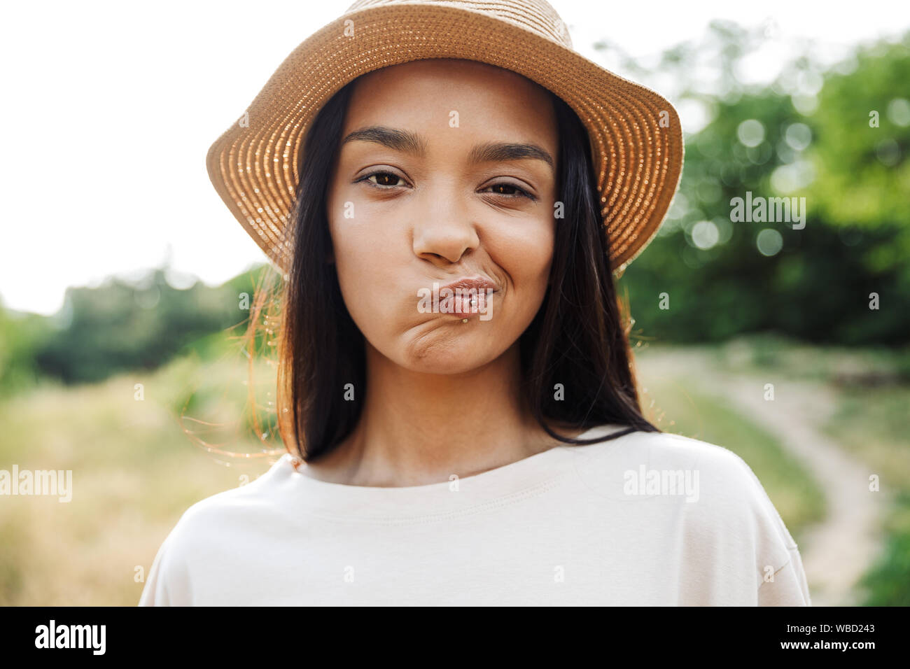 Photo of caucasian woman wearing straw hat and lip piercing twisting her mouth while walking in green park Stock Photo