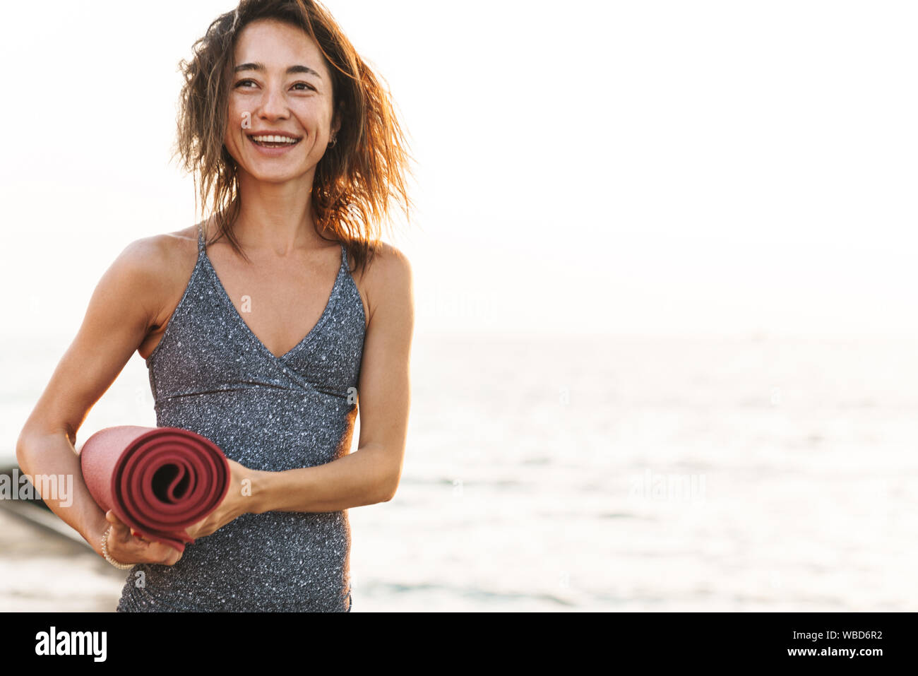 Photo of brunette sportswoman in tracksuit smiling and standing with yoga mat by seaside in morning Stock Photo