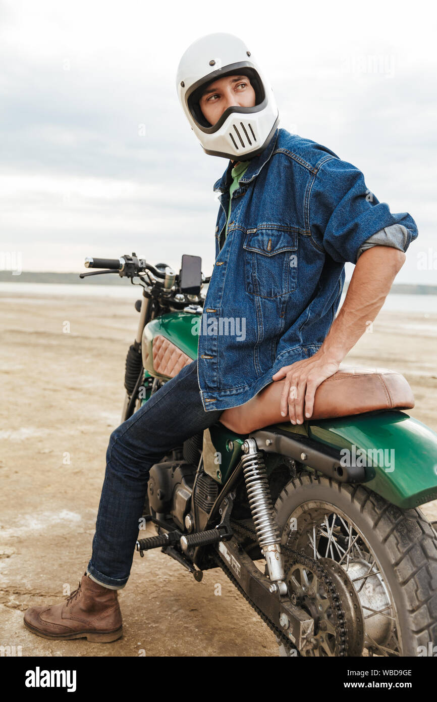 Handsome young man wearing casual outfit sitting on a motocycle at the beach, wearing a helmet Stock Photo
