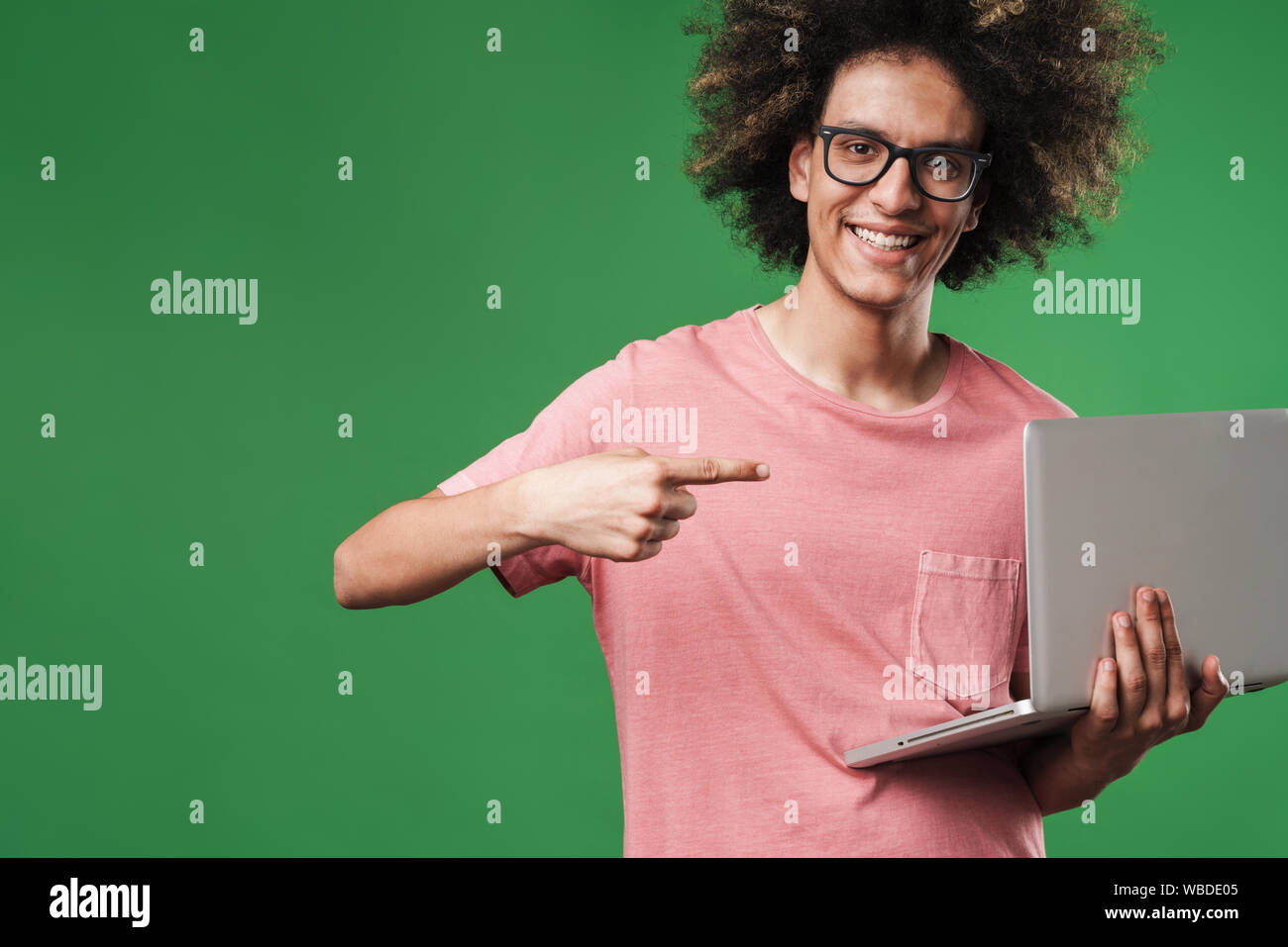 Photo of a young positive happy curly guy posing isolated over green wall background using laptop computer pointing to copyspace. Stock Photo