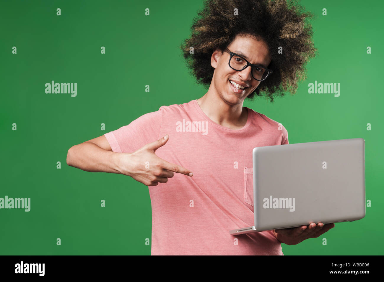 Photo of a young positive smiling cheery curly guy posing isolated over green wall background using laptop computer pointing to copyspace. Stock Photo