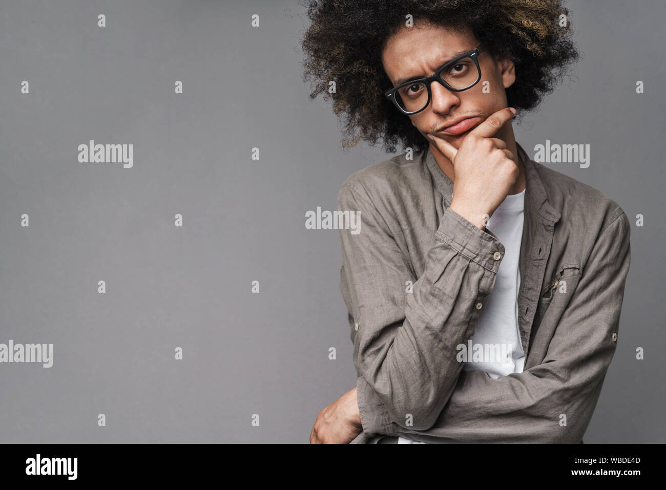 Photo of brooding unshaven man with afro hairstyle propping up his head and looking at camera isolated over green background Stock Photo