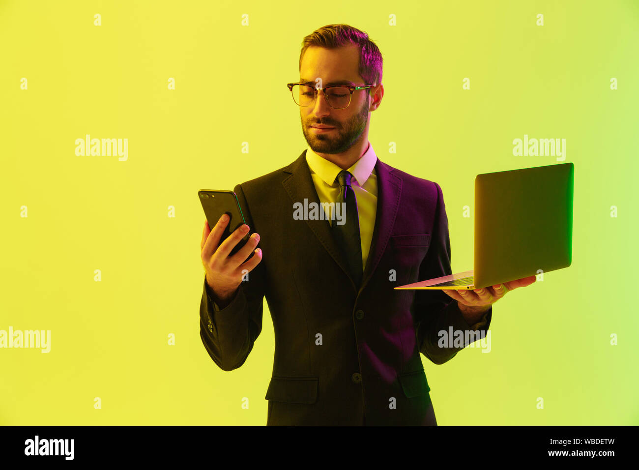 Image of a handsome young business man posing isolated over light green background wall with led neon lights using laptop computer using mobile phone. Stock Photo