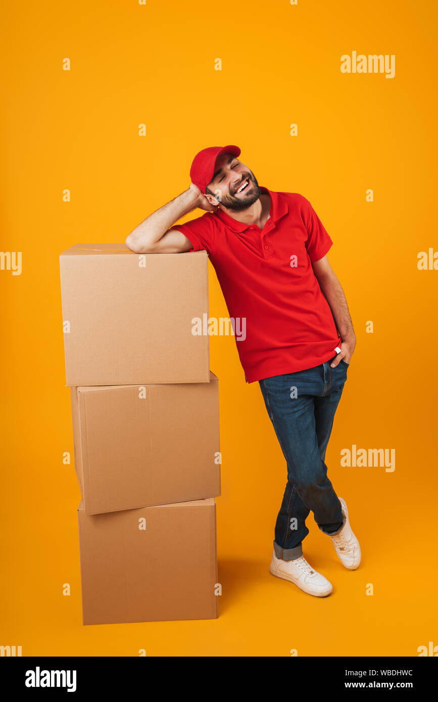 Portrait of happy delivery man in red uniform smiling while standing with packaging boxes isolated over yellow background Stock Photo