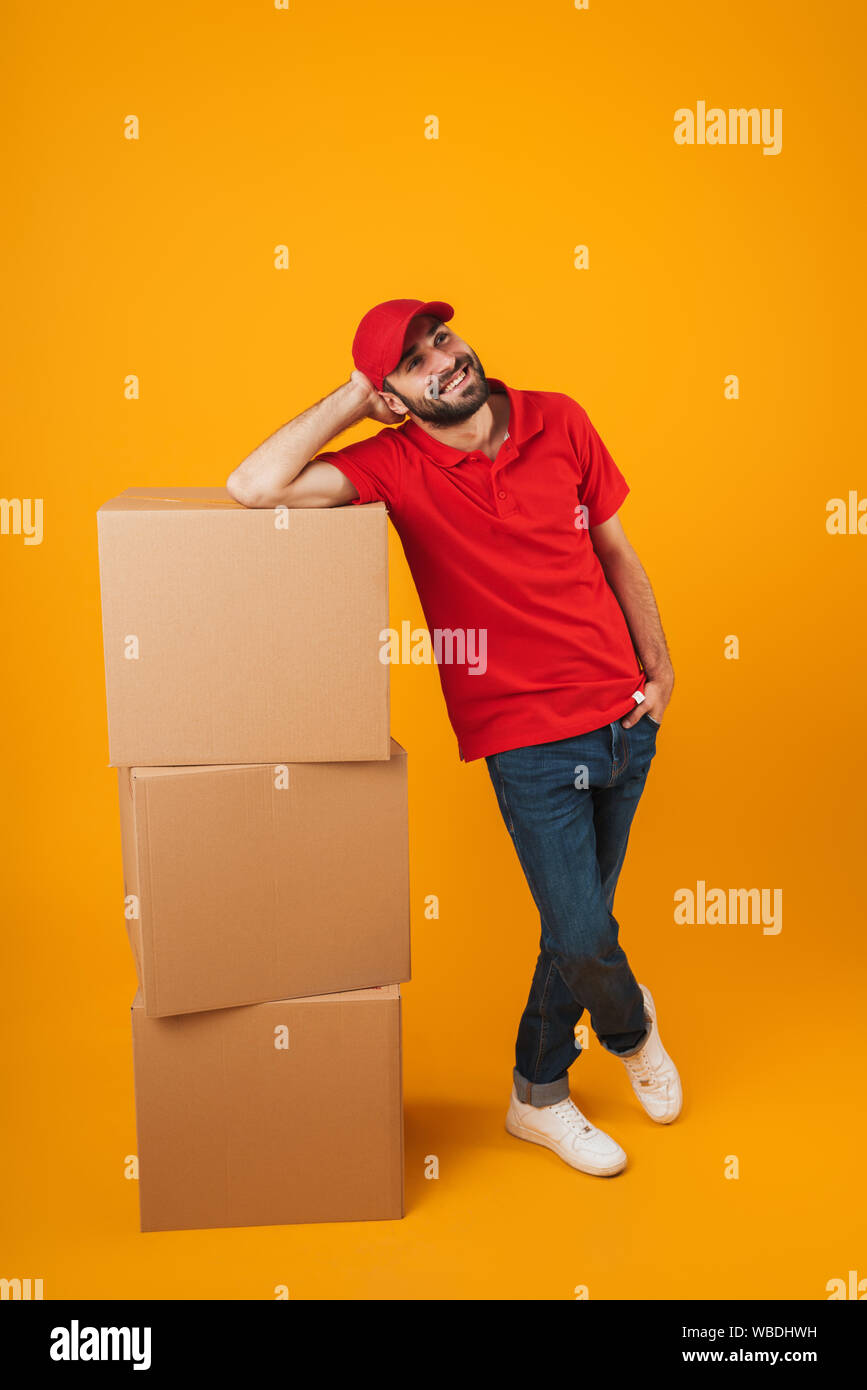 Portrait of cheerful delivery man in red uniform smiling while standing with packaging boxes isolated over yellow background Stock Photo