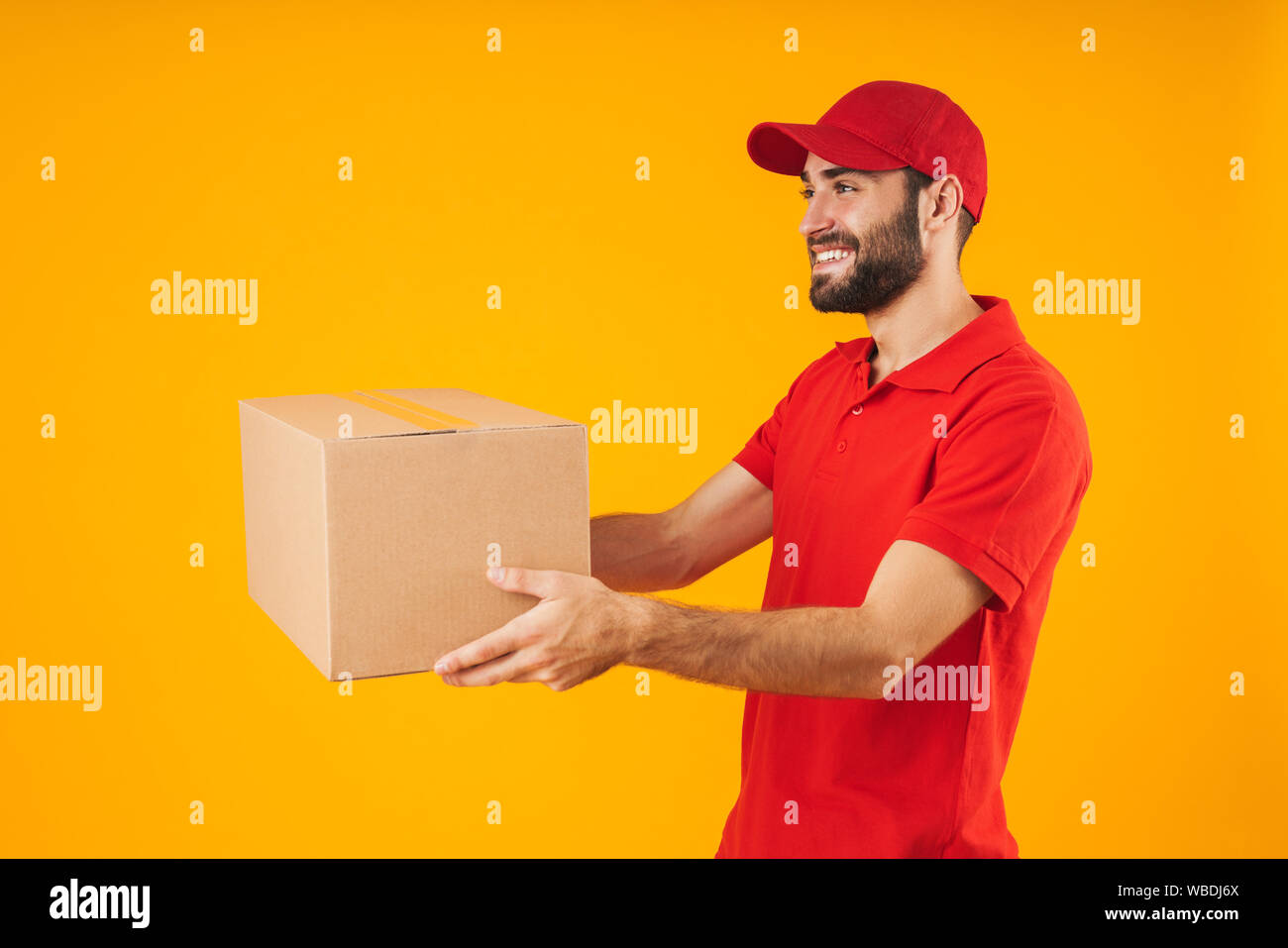 Image of pleased delivery man in red uniform smiling and holding packaging box isolated over yellow background Stock Photo