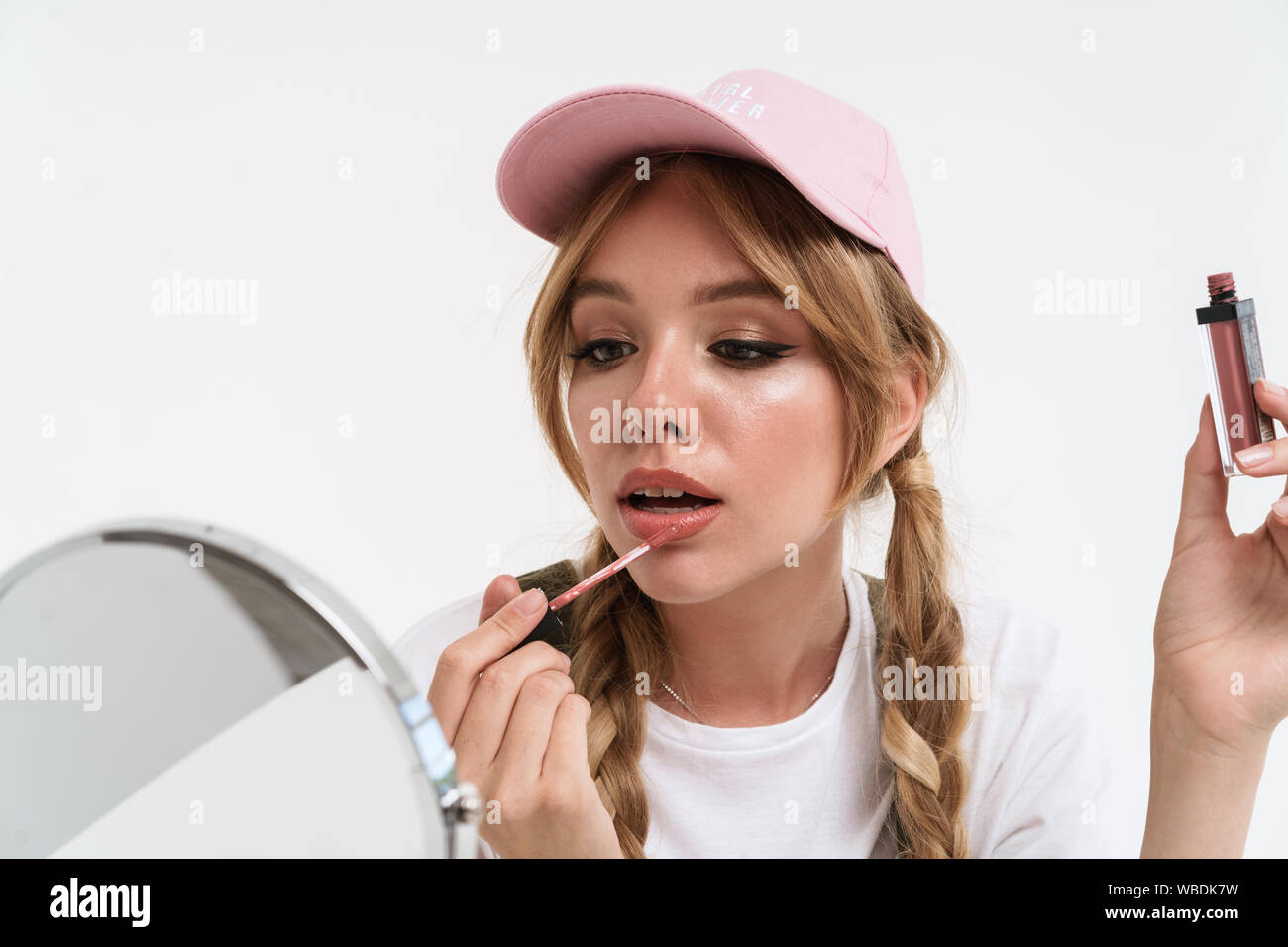 Close up of a beautiful young girl wearing cap doing makeup while looking at the mirror isolated over white Stock Photo