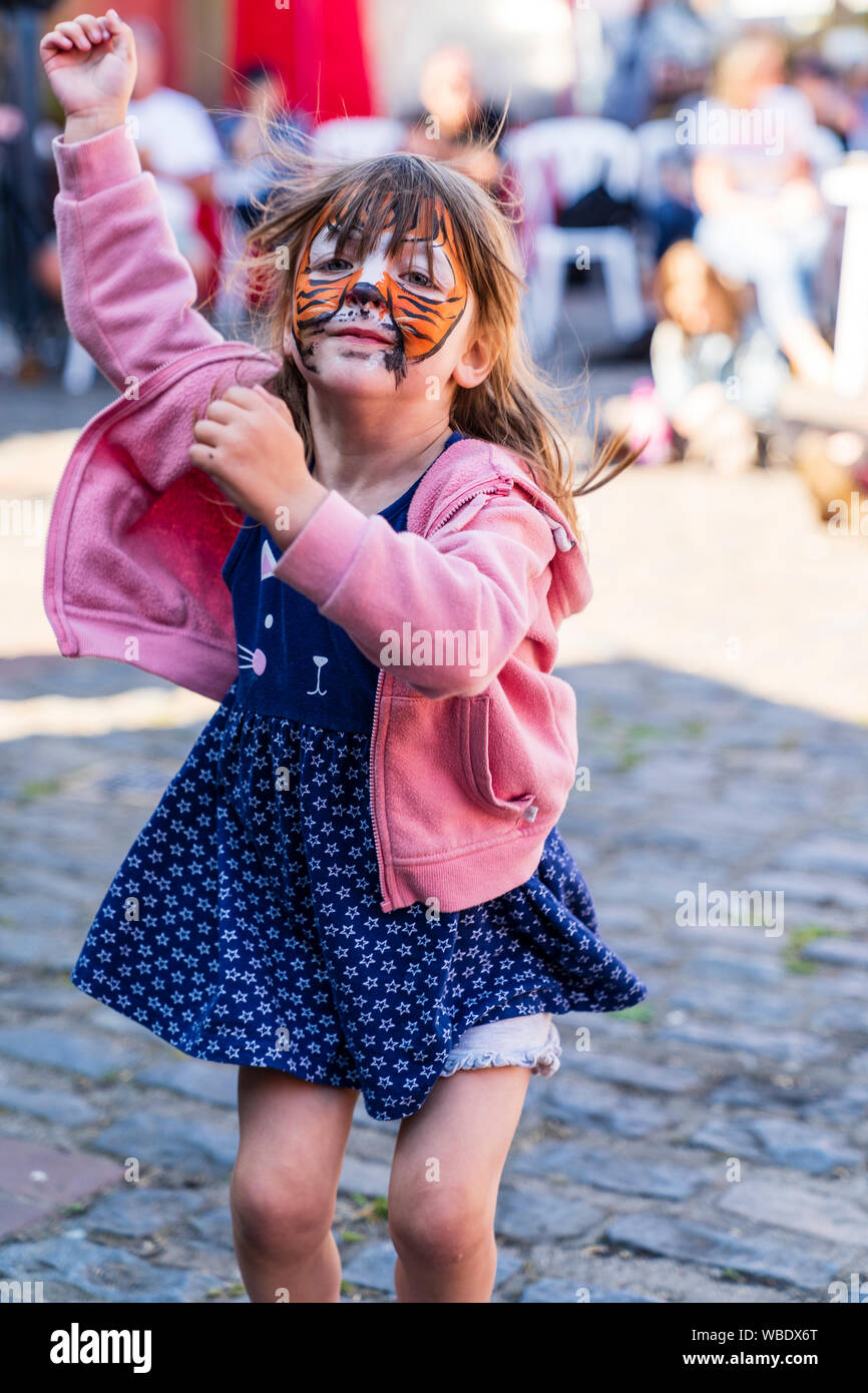 Caucasian child, girl, 5-7 year old, with painted tiger face, dancing by jumping up and down during outdoor musical event. Facing viewer, eye-contact. Stock Photo