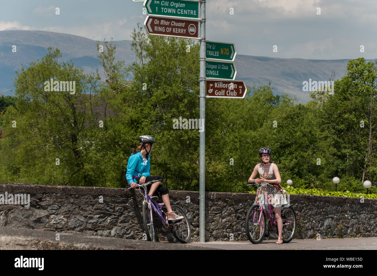 Two 2 young women tourists with bicycles wondering about the direction at a road junction. Road signs in English and Irish language writing. Stock Photo