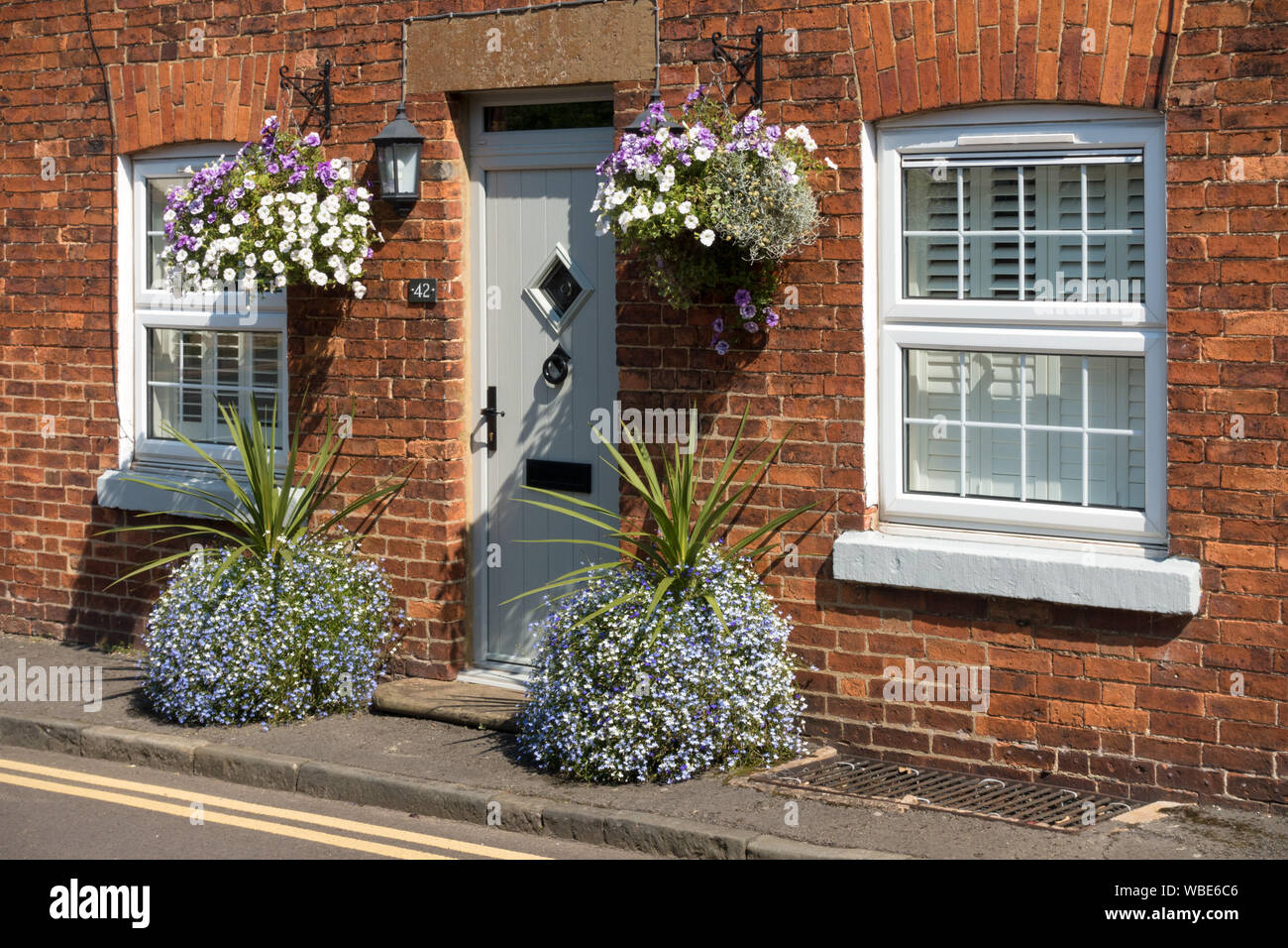 Old red brick cottage with modern UPVC double glazed windows and front door with pretty flowers in hanging baskets and pots, Uppingham, Rutland, UK. Stock Photo
