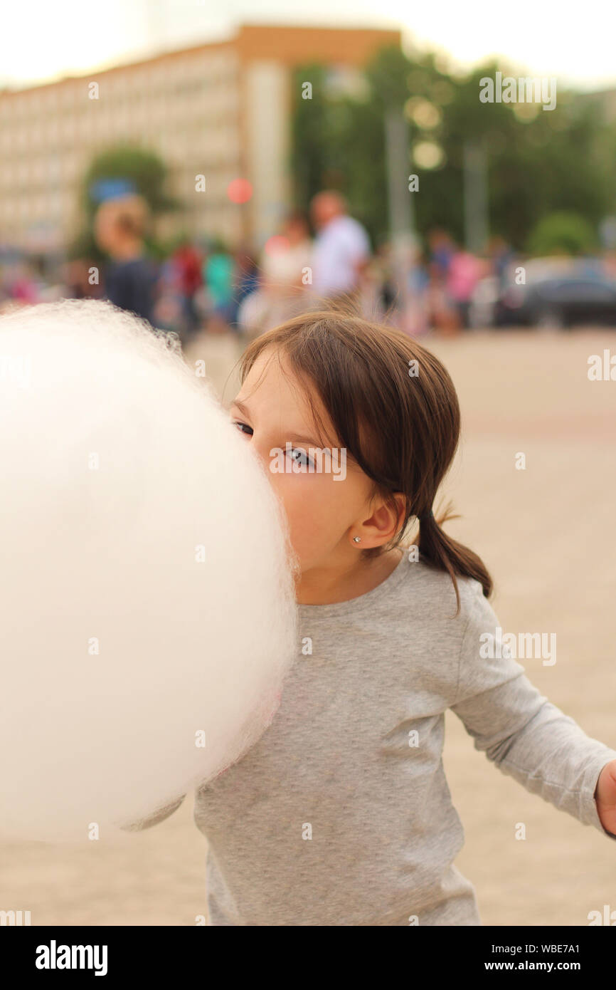 Little girl with pigtails tries to bite a big ball of cotton candy at a city celebration Stock