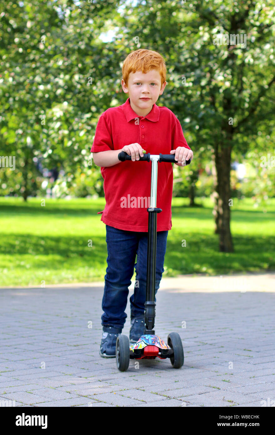 Child learn to ride scooter in a city park on sunny summer day. Cheerful little boy with ginger hair posing with a scooter. Active leisure and outdoor Stock Photo