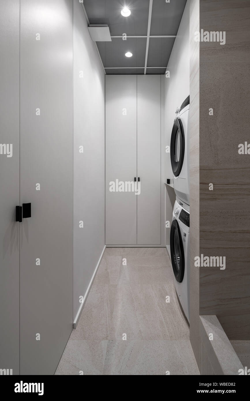 Contemporary laundry room with textured beige tiled walls and luminous lamps. There are light lockers, two washing machines which stand on each other. Stock Photo