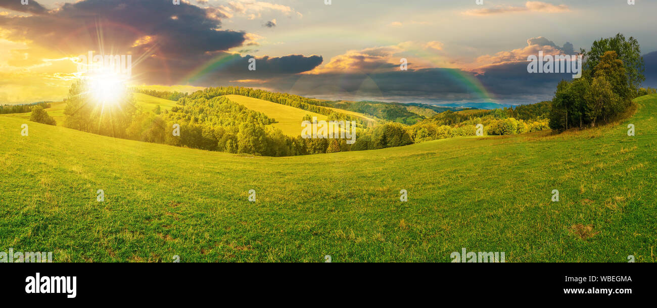 panoramic mountain landscape at sunset. grassy meadow on the hillside in evening light. trees on the edge of a hill. mountain ridge in the distance. c Stock Photo
