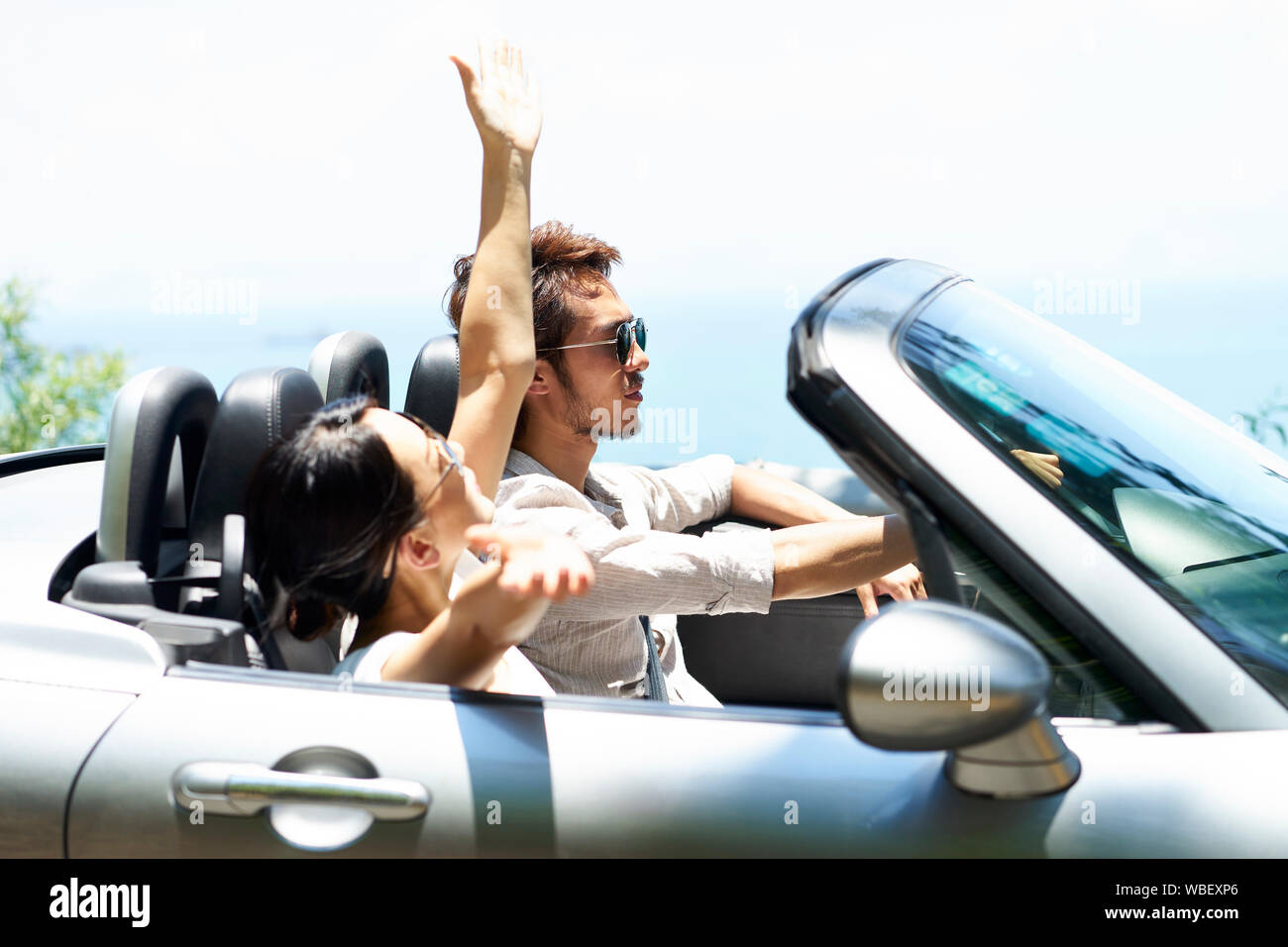 young asian couple riding in a convertible car on a seaside road, side view Stock Photo