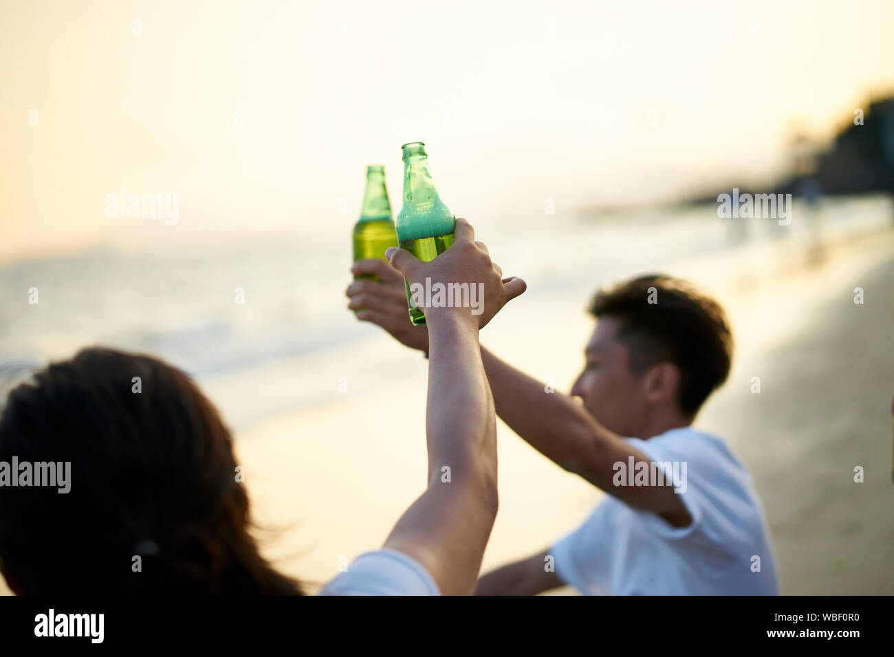 two young asian adult men drinking beer clicking bottles toasting on beach Stock Photo