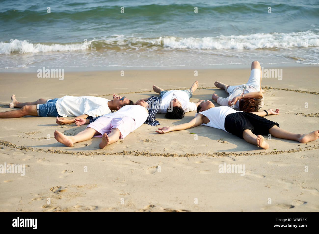 group of five young asian adults men lying relaxed on sand beach forming a circle Stock Photo