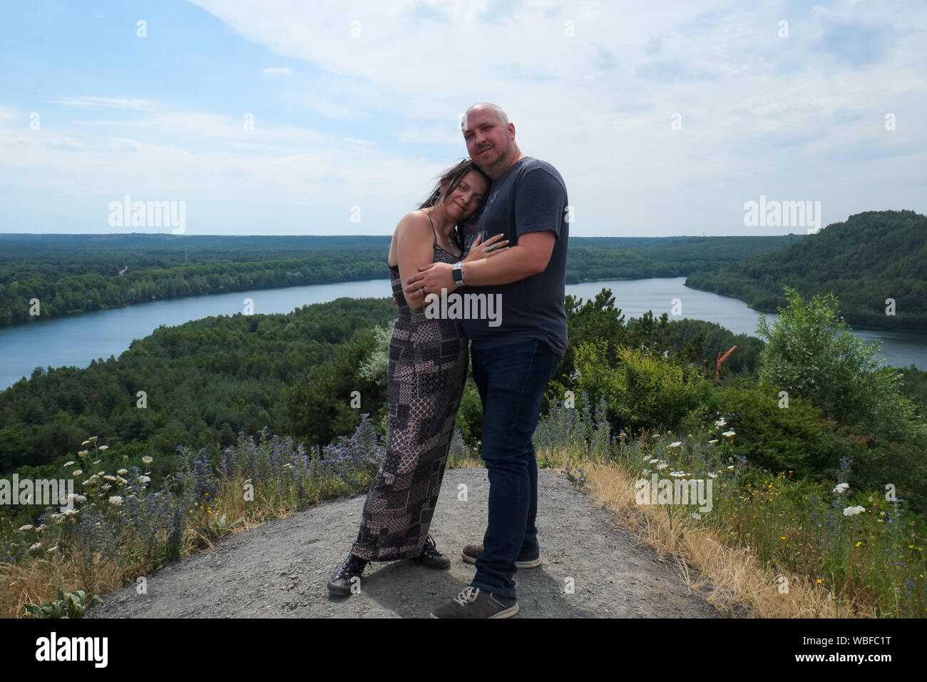 Loving couple of 30-40 years old outdoors hugging and standing tall on a hill with a river running through the woods the background Stock Photo