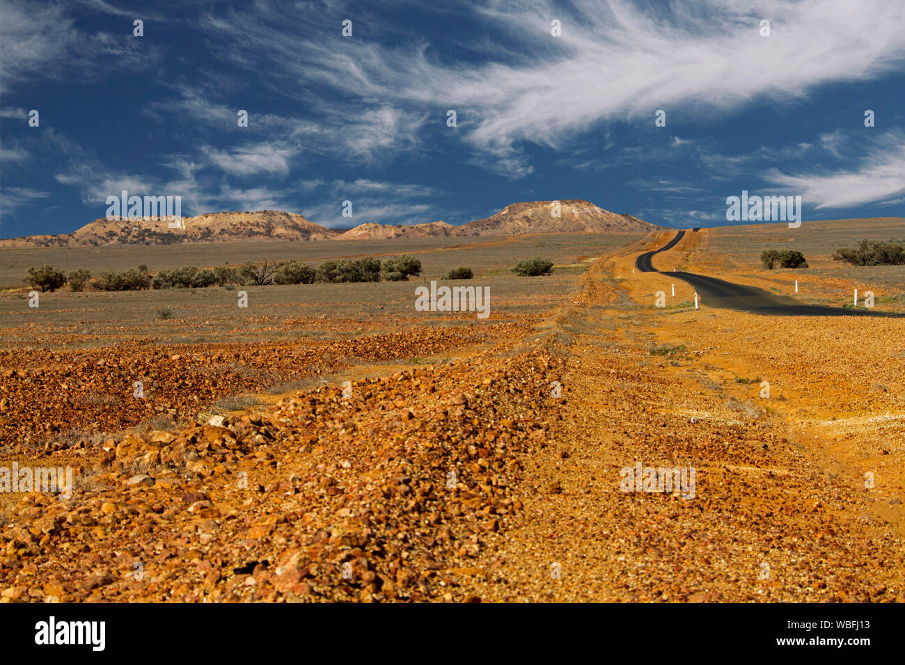 Road stretching across stony barren plains that stretch to distant hills and horizon under blue sky in outback Queensland during drought Stock Photo