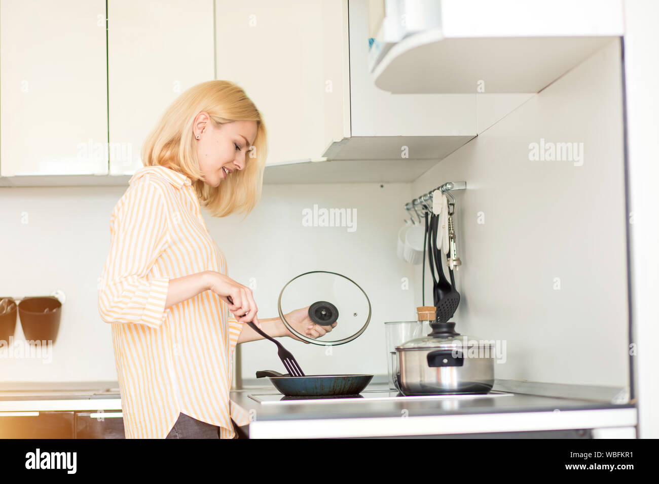 Happy beautiful caucasian blonde woman standing in kitchen near stove with spatula, cooking eggs in frying pan in morning. Female student cooking brea Stock Photo