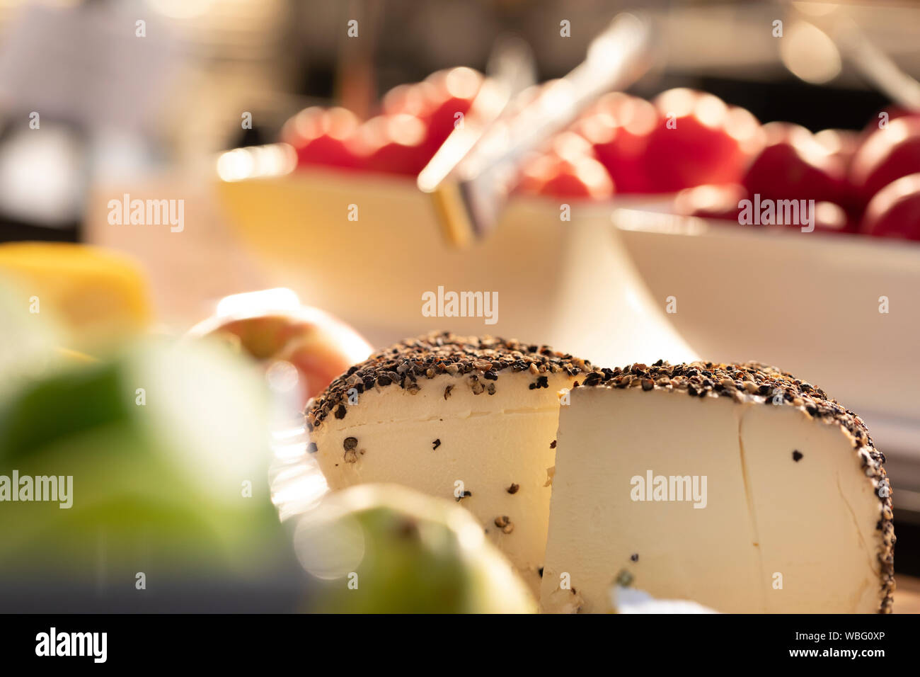 slice of cheese with a pepper rind on a buffet and blurred tomatoes in the background Stock Photo