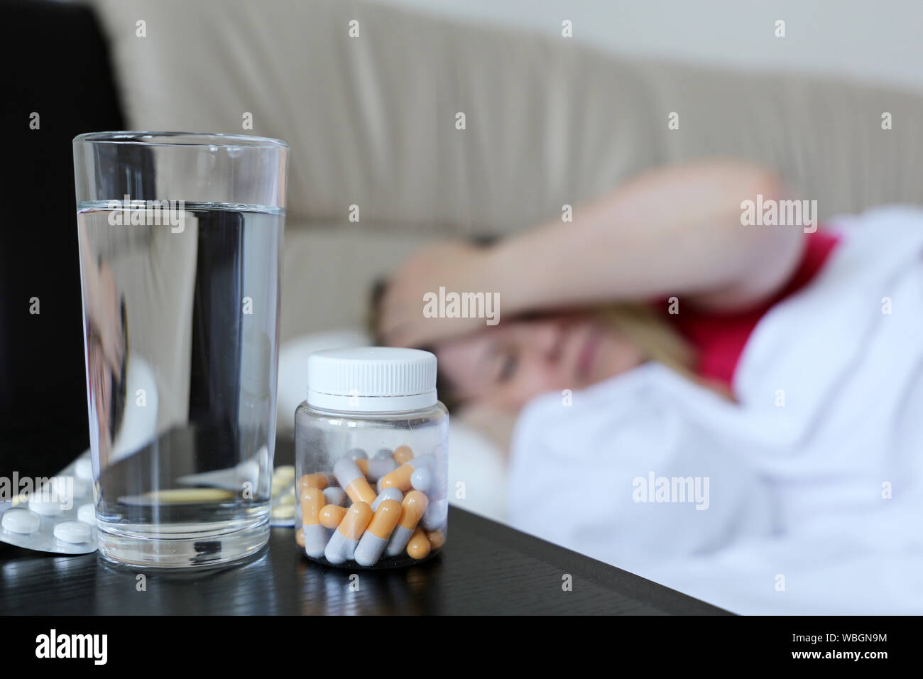 Sick woman lies in a bed holding hand to her forehead, cold and flu season. Pills and water glass in the foreground, concept of illness, fever Stock Photo