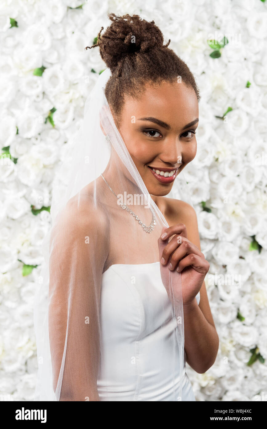 Happy African American Bride Touching White Veil Near Flowers