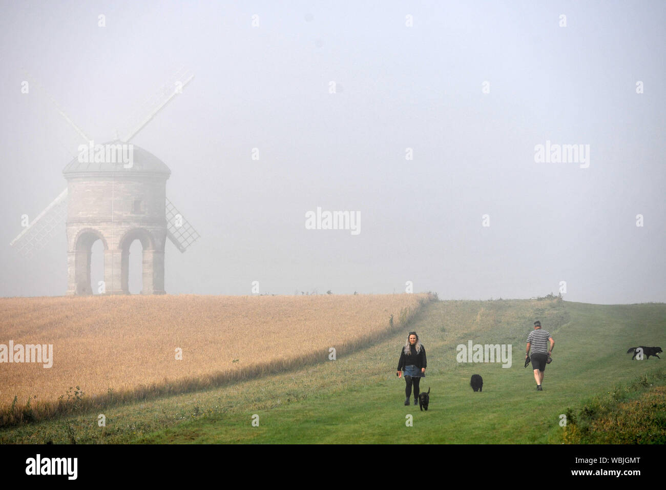 People walk their dogs through a layer of morning mist by Chesterton Windmill, Warwickshire, following the hottest August Bank Holiday Monday on record, with temperatures reaching 33.2C. Despite the mist, the heatwave is set to continue for some parts of the UK after the record-breaking bank holiday weather. Stock Photo